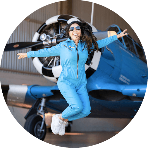 Cláudia Ruete usando uma roupa azul, enquanto salta sorrindo em frente a um avião azul ao fundo